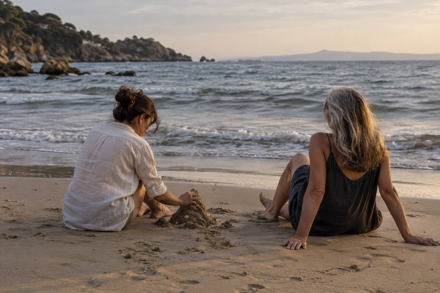 Dos mujeres sentadas junto al mar, con los pies sobre la arena y pequeñas construcciones de arena entre ellas.